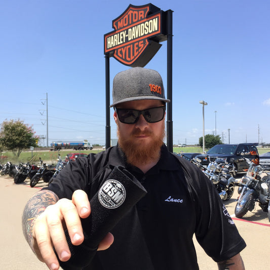 Man in a black shirt and cap standing in front of a Harley-Davidson sign with motorcycles in the background.
