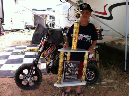 A kid holding a trophie with his dirt bike in the background with red Shock Sox on the bike.