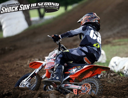 A person on a dirt bike with Shock Sox branding on the handlebar, wearing a helmet and numbered suit, with the bike in an off-road racing setting.
