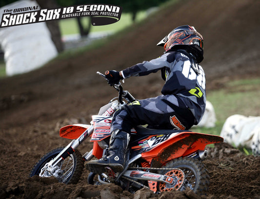 A person on a dirt bike with Shock Sox branding on the handlebar, wearing a helmet and numbered suit, with the bike in an off-road racing setting.