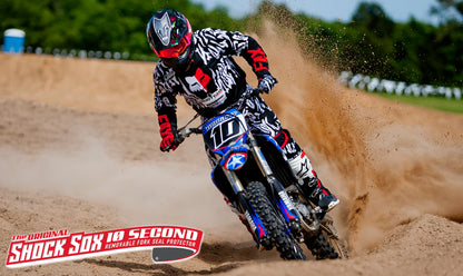 A motocross rider wearing a checkered helmet and riding a dirt bike, with the 6" DIRT SHOCK SOX branding visible on the bike's fork.