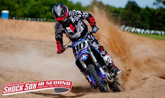A motocross rider wearing a checkered helmet and riding a dirt bike, with the 6" DIRT SHOCK SOX branding visible on the bike's fork.